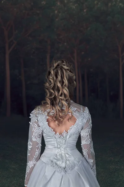 Back view of a woman in white wedding dress standing in moody dark forest. Rear view