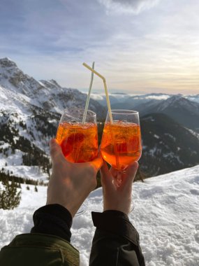 Bir çift, bir dağın tepesinde pipetli cam kadehlerde Aperol tutuyor. Dağların ve karın panoramik manzarası. Dağdaki aşıkların elleri.