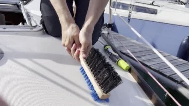 Bir kişi, yat limanının yakınındaki bir yatın güvertesini titizlikle ovalıyor. Detaylı video çekimleri için fırça kullanıyor.