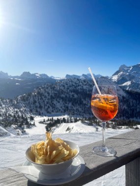 Serinletici bir deneyim için açık mavi gökyüzünün altındaki karlı bir dağın tepesinde Aperol Spritz yudumla ve cips ye.