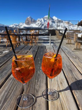 İki Aperol Spritz kokteyli, ahşap bir masanın üzerinde. Karlı bir dağ manzarası var.