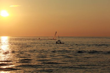 Suda süzülen bir yelkenlinin yer aldığı sakin ve resimli bir gün batımı sahnesi.