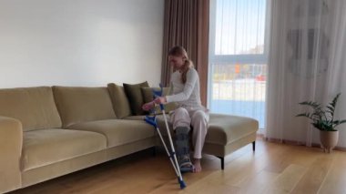 Bacağı alçılı bir kadın oturma odasındaki kanepeden kalkar ve yavaşça ileri doğru yürür..