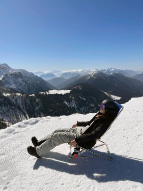 Karlı bir dağdaki bir sandalyede rahatça dinlenen bir snowboardcu, nefes kesici manzaranın ve açık mavi gökyüzünün tadını çıkarıyor, kış manzarasının huzurlu atmosferini kucaklıyor..