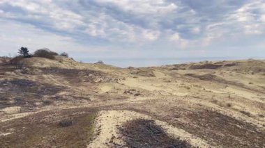 Sakin ve sakin bir manzara bulutlu bir gökyüzünün altında sakin denize doğru uzanan kumulları sergiliyor.