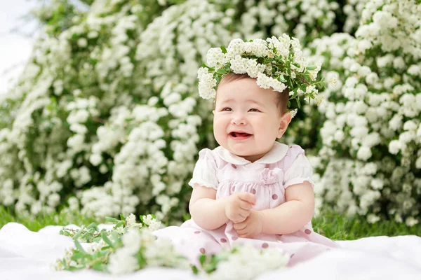 Sweet smiling little girl with wreath of spirea flowers in pink dress, on green grass near spirea bushes on sunny summer day closeup