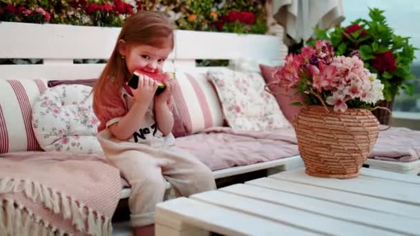 小さな赤ん坊の女の子は家で夏のパティオに座っている間にスイカを食べて — ストック動画