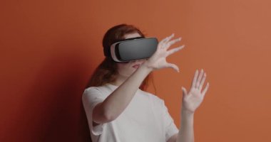Kızıl saçlı kadın sanal gerçekliğin tadını çıkarıyor. VR kulaklık takan, elleriyle havaya dokunan, heyecanlı ifadeyle sanal oyun oynayan bir kızın portresi. Turuncu arka planda kapalı stüdyo çekimleri