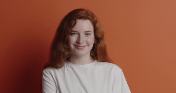赤毛の女がカメラをポーズ 正の女の子のトレーナーを着て誠実に笑みを浮かべて カメラを見て いちゃつく オレンジの背景に隔離された屋内スタジオショット — ストック動画