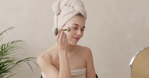 フェイスマッサージ 肌のケア 自宅での美しさの治療のための玉顔ローラーを使用してバスローブの女性 鏡を見てシャワー後マッサージをする女の子 — ストック動画