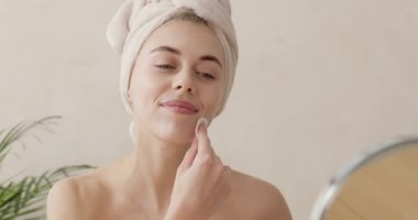 Bathrobe woman cleaning face with white pad. Young woman caring for facial skin using cotton pad and looking in round mirror in bathroom interior. Beauty care and pampering concept 