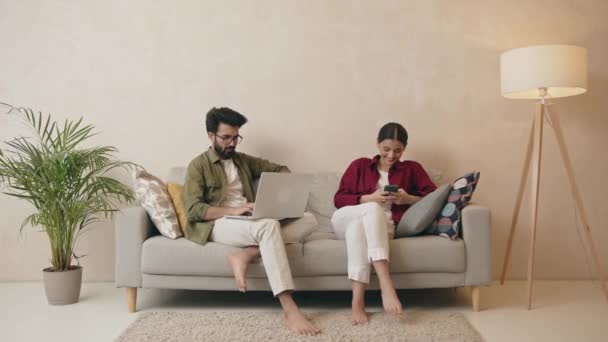 幸せなインドのカップルのリビングルームでソファに座って ノートパソコンやスマートフォンを使用しています 人々は家で時間を過ごす 家庭生活とライフスタイルの概念 — ストック動画