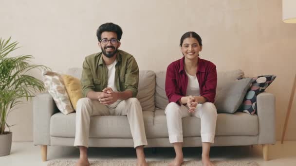 若い髭を生やした男と魅力的な喜びの妻がソファに座って カメラを見て微笑んだ 幸せなインドの家族カップル見てコメディアン映画面白いビデオ 楽しい休日の週末時間一緒に家で — ストック動画