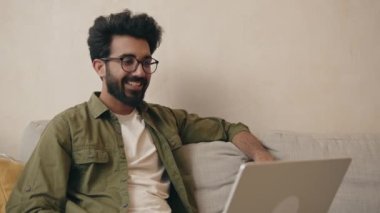 Mutlu Hintli adam diz üstü bilgisayarla ararken selam veriyor oturma odasındaki kanepede oturuyor. Çevrimiçi iletişim kuran neşeli adam, evdeki modern aletleri kullanıyor. 