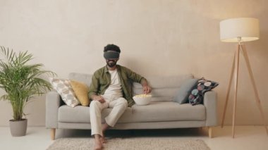 Heyecanlı adam VR gözlükleri. Sanal gerçeklik cihazı takan genç adam patlamış mısır yiyor ve kanepede otururken film izliyor. Yeni trendler teknoloji kavramı 