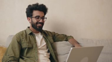 Mutlu Hintli adam diz üstü bilgisayarla ararken selam veriyor oturma odasındaki kanepede oturuyor. Çevrimiçi iletişim kuran neşeli adam, evdeki modern aletleri kullanıyor. 