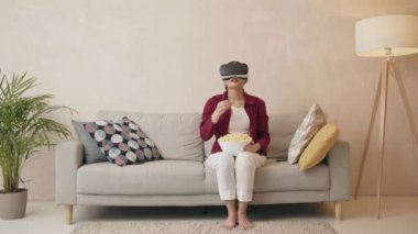 Excited woman VR glasses. Young girl wearing virtual reality device eating popcorn and watching movie while sitting on sofa. New trends technology concept 