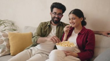 Genç mutlu Hintli çift TV 'de film izliyor ve oturma odasında dinlenirken patlamış mısır yiyor. 