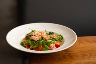 White ceramic plate of fresh salad with salmon, vegetables, tomatoes and arugula with sesame seeds.