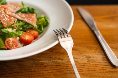 White ceramic plate of fresh salad with salmon, vegetables, tomatoes and arugula with sesame seeds.