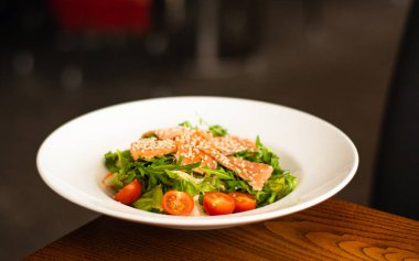 White ceramic plate of fresh salad with salmon, vegetables, tomatoes and arugula with sesame seeds.