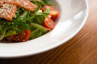 White ceramic plate of fresh salad with salmon, vegetables, tomatoes and arugula with sesame seeds.