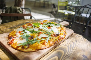 Sebze ve peynirle kaplı yeni pişmiş bir pizza güneşli bir günde, sıcacık bir kafede, ahşap bir tahtanın üzerinde oturur.