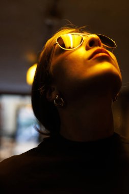 Yansıtıcı güneş gözlüğü takan genç bir kadın sıcak bir şehir kafe ortamında gece boyunca yukarı bakıyor.