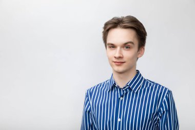 Portrait of confident and cheerful young man wearing blue-white striped button shirt looking at camera with one eyebrow raised. Studio shot on gray background