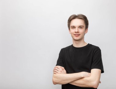 Portrait of cheerful young man wearing black tshirt standing with arms crossed smiling at camera. Studio shot on gray background