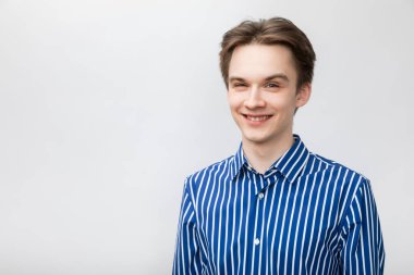 Portrait of happy cheerful teenager boy wearing blue-white striped button shirt looking at camera smiling. Studio shot on gray background