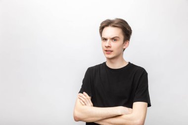 Portrait of doubtful young man wearing black tshirt holding his hands crossed looking at camera suspiciously. Studio shot on gray background