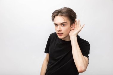 Portrait of young man wearing black tshirt listening holding hand near ear looking at camera. Studio shot on gray background