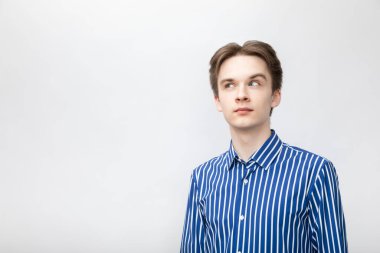 Portrait of thoughtful young man wearing blue-white striped button shirt looking side. Studio shot on gray background