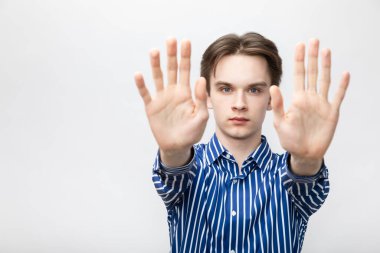 Portrait of confident young man wearing blue-white striped button shirt holding out his hands showing stop gesture looking at camera. Studio shot on gray background