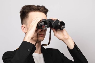 Portrait of elegant young man wearing black  suit looking through binoculars. Studio shot on gray background