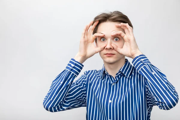 Portrait of teenager boy wearing blue-white striped button shirt holding his eyes wide open with fingers. Studio shot on gray background
