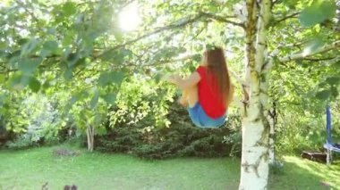 Kırmızı tişört giyen mutlu bir kız ağaçtan aşağı sarkıyor ve güneşli bir bahçede oynuyor. Yaz tatilinin tadını çıkarıyor.