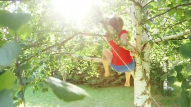 Kırmızı tişört giyen mutlu bir kız ağaçtan aşağı sarkıyor ve güneşli bir bahçede oynuyor. Yaz tatilinin tadını çıkarıyor.