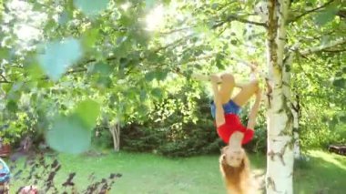 Kırmızı tişört giyen mutlu bir kız ağaçtan aşağı sarkıyor ve güneşli bir bahçede oynuyor. Yaz tatilinin tadını çıkarıyor.
