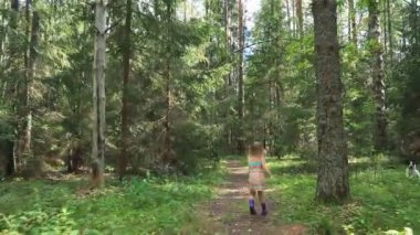 Yazlık elbiseler ve lastik botlar giyen küçük bir kız sıcak yaz gününde ormanda yol boyunca yürüyor. 
