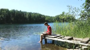 Orman gölünün kıyısındaki ahşap iskelede oturan genç çocuk pastel boya kalemleriyle resim çiziyor.