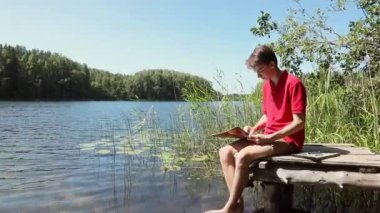 Orman gölünün kıyısındaki ahşap iskelede oturan genç çocuk pastel boya kalemleriyle resim çiziyor.