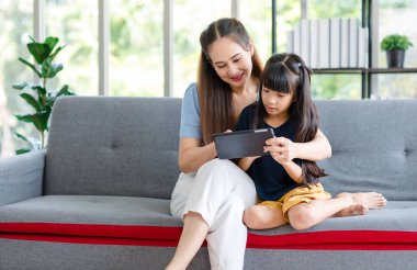 Milenyum Asyalı mutlu aile annesi rahat koltukta oturmuş gülümsüyor ve dokunmatik ekranlı akıllı telefondan evdeki küçük kız çocuğuna komik içerik yayını yapıyor..