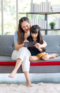 Milenyum Asyalı mutlu aile annesi rahat koltukta oturmuş gülümsüyor ve dokunmatik ekranlı akıllı telefondan evdeki küçük kız çocuğuna komik içerik yayını yapıyor..