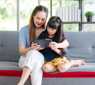 Milenyum Asyalı mutlu aile annesi rahat koltukta oturmuş gülümsüyor ve dokunmatik ekranlı akıllı telefondan evdeki küçük kız çocuğuna komik içerik yayını yapıyor..