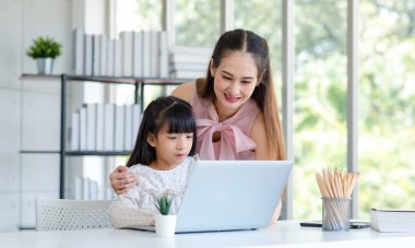 Milenyum Asyalı mutlu aile annesi gülümsüyor. Küçük kız çocuğuna öğretmenlik yapmasına yardımcı oluyor. Evdeki oturma odasında dizüstü bilgisayarla okul ödevi yapıyor..