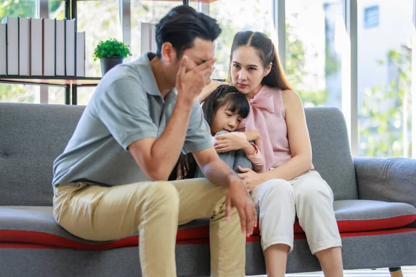 Millennial Asian unhappy family mother sitting on sofa couch holding hugging soothing little young female daughter while sad stressed father crying after big fight argument in living room at home.