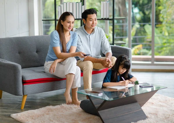 Millennial Asian happy family father and mother sitting on cozy sofa couch holding remote watching television while little young girl daughter sitting on floor doing homework in living room at home.