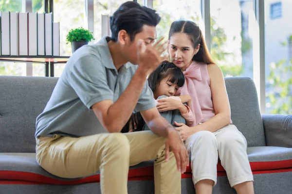 Millennial Asian unhappy family mother sitting on sofa couch holding hugging soothing little young female daughter while sad stressed father crying after big fight argument in living room at home.